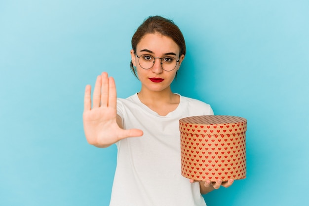 Jeune fille arabe maigre tenant une boîte de jour de valentines debout avec la main tendue montrant le panneau d'arrêt, vous empêchant.