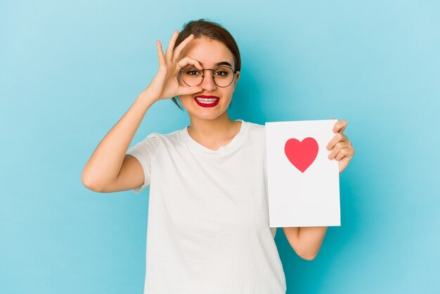 Jeune fille arabe maigre tenant une carte de Saint Valentin excitée en gardant le geste ok sur les yeux.