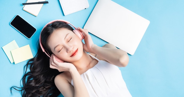 Jeune fille asiatique couchée sur bleu avec ordinateur portable, smartphone, écouteurs et note autour de bleu