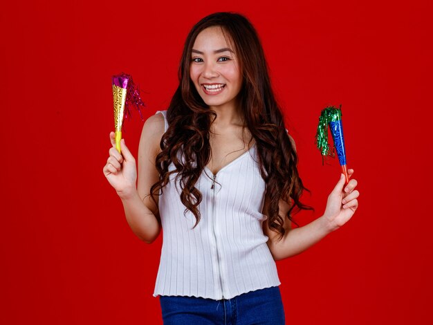 Jeune fille asiatique drôle et guérie tenant et prête à souffler en jouant avec des popers drôles et heureux. Studio tourné sur fond rouge.