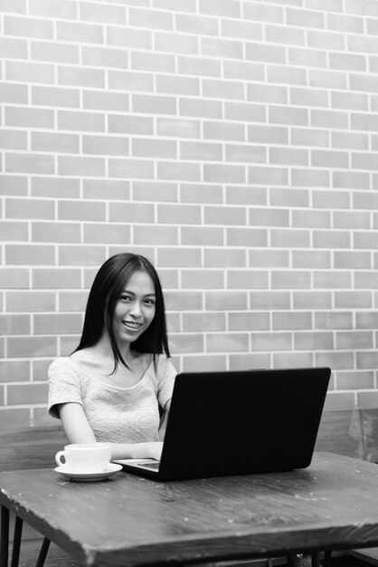 Jeune fille asiatique heureuse souriant tout en utilisant un ordinateur portable avec cappuccino sur une table en bois sur un mur de briques