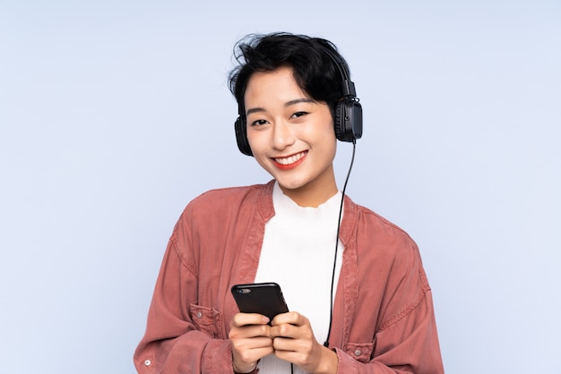 Jeune fille asiatique sur mur bleu isolé écouter de la musique avec un mobile et à l'avant