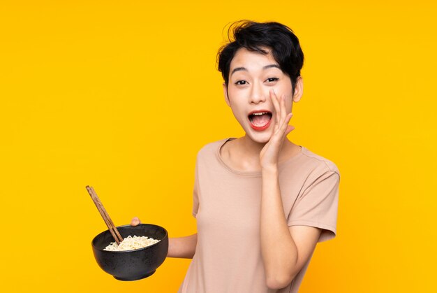 Jeune fille asiatique sur mur jaune isolé criant avec la bouche grande ouverte tout en tenant un bol de nouilles avec des baguettes