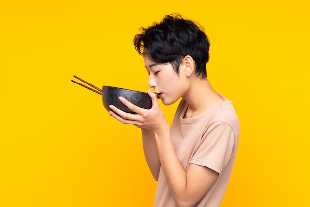 Jeune fille asiatique sur mur jaune isolé tenant un bol de nouilles avec des baguettes et le manger