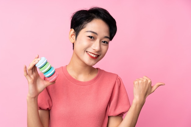 Jeune fille asiatique sur un mur rose isolé tenant des macarons français colorés et pointant vers le côté