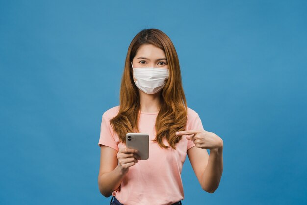 Jeune fille asiatique portant un masque médical utilisant un téléphone portable avec des vêtements décontractés isolés sur un mur bleu