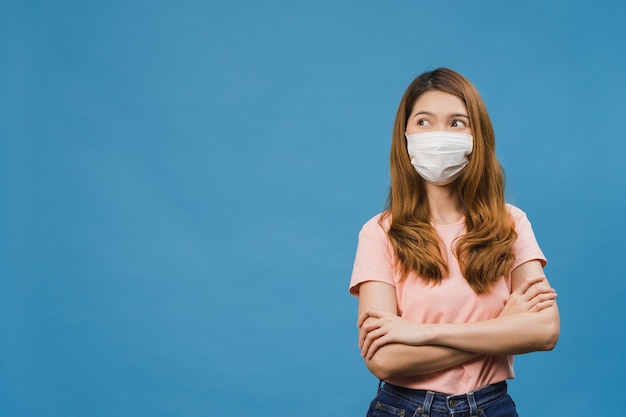 Jeune fille asiatique portant un masque médical vêtu d'un tissu décontracté et regardant un espace vide isolé sur fond bleu