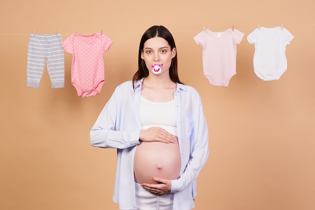 Une jeune fille assez enceinte, anxieuse et fatiguée, avec une tétine dans la bouche, tient doucement ses mains sur son ventre, regarde droit, se tient entourée de vêtements pour enfants séchant sur une corde.