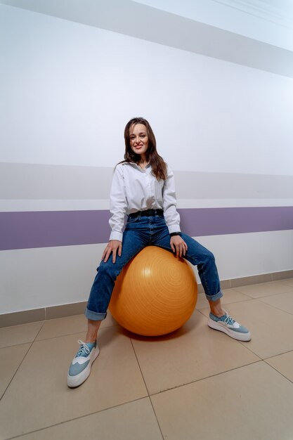 Jeune fille assise sur un ballon de fitness orange. Une femme en jeans apprend des exercices avec un ballon.