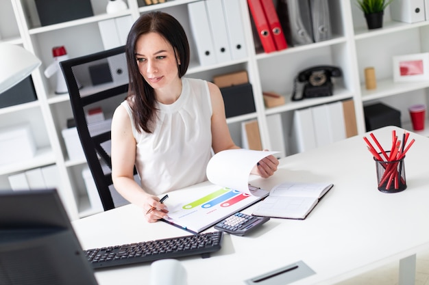 Une jeune fille assise dans le bureau à un bureau d'ordinateur et travaillant avec des documents et une calculatrice.