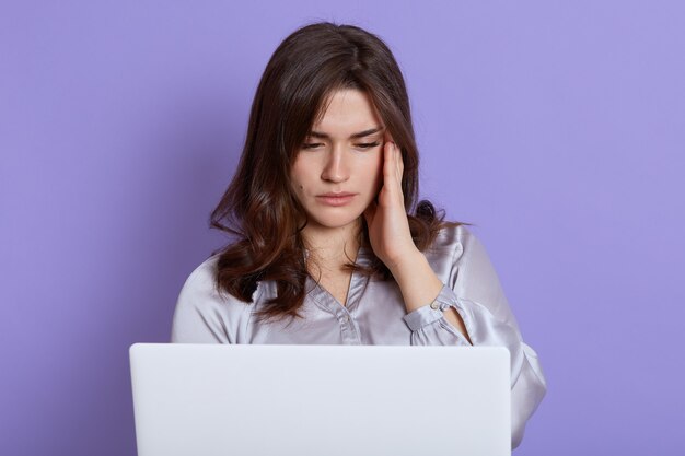 Jeune fille assise devant un ordinateur portable blanc avec une expression faciale épuisée