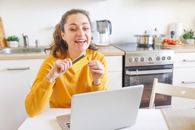 Jeune fille assise avec un ordinateur portable acheter sur Internet entrer les détails de la carte de crédit sur l'intérieur de la cuisine