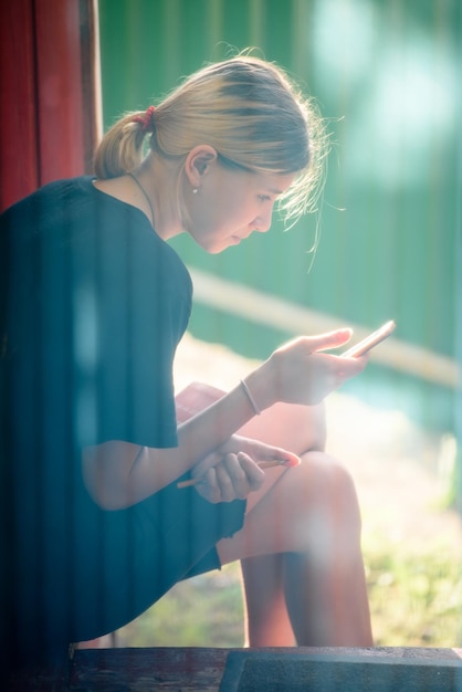 Jeune fille assise sur le porche d'une maison dans une maison chaude et regardant dans le smartphone