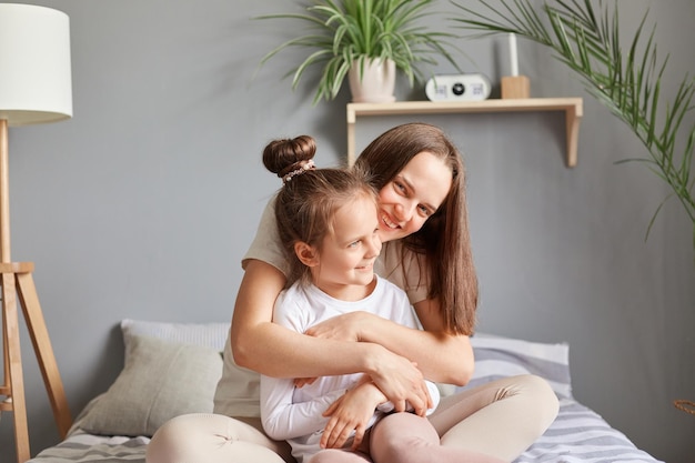 Jeune fille assise avec sa mère dans leur chambre confortable souriante et se sentant ensemble dans une relation profitant d'une journée tranquille remplie d'amour et de détente