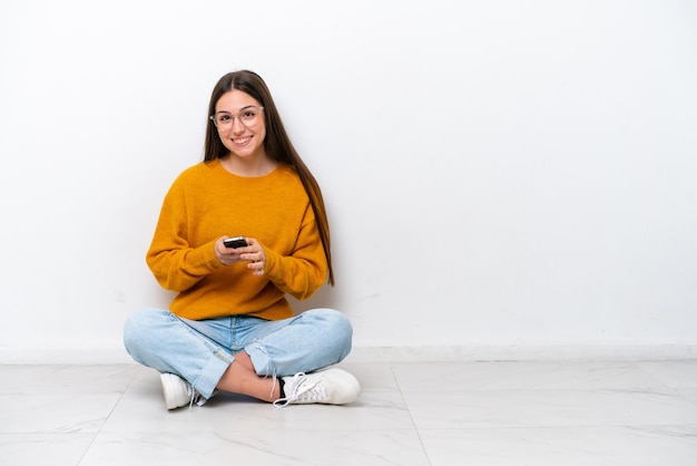 Jeune fille assise sur le sol isolé sur fond blanc en envoyant un message avec le mobile