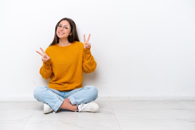 Jeune fille assise sur le sol isolé sur fond blanc montrant le signe de la victoire avec les deux mains