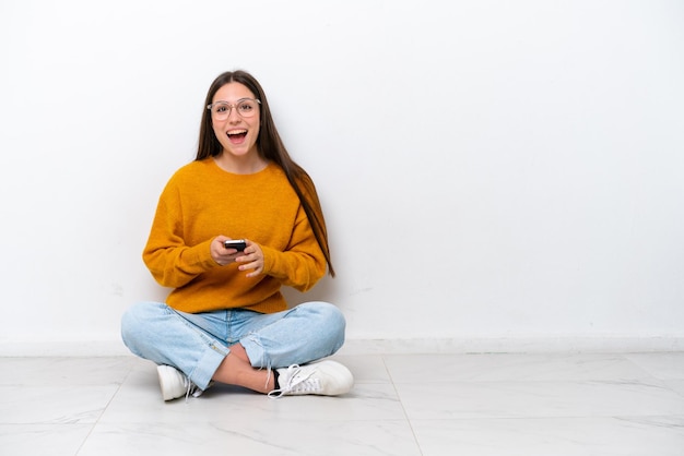 Jeune fille assise sur le sol isolé sur fond blanc surpris et envoyant un message