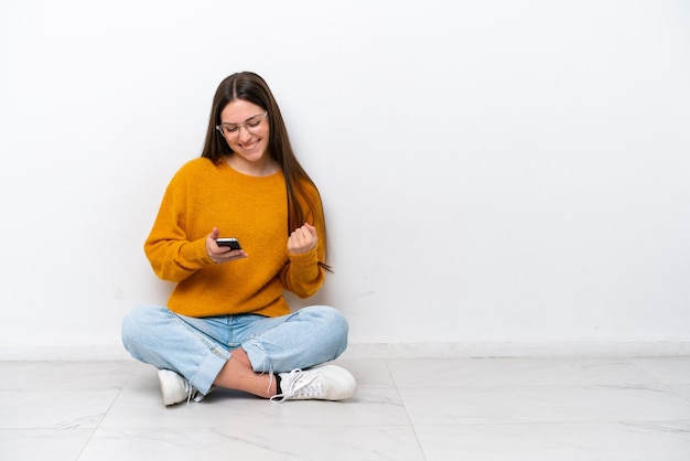 Jeune fille assise sur le sol isolé sur fond blanc avec téléphone en position de victoire