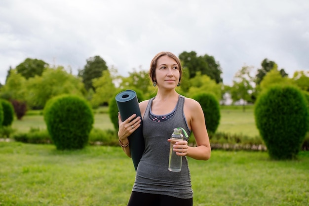 Une jeune fille athlétique en survêtement gris pour le fitness va faire du yoga dans un parc verdoyant, de l'eau.