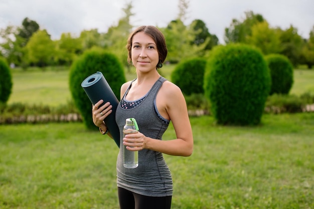 Une jeune fille athlétique en survêtement gris pour le fitness va faire du yoga dans un parc verdoyant.