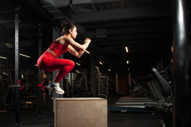 Jeune fille athlétique en tenue de sport rouge sautant dans une salle de sport noire jolie femme faisant du sport