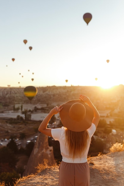 La jeune fille attirante se tient sur la montagne avec des ballons à air volants sur le fond Cappadocia