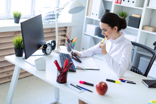 Une jeune fille au bureau a un stylo dans la bouche et travaille avec une calculatrice, des documents et un ordinateur.