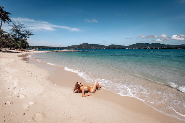 Une jeune fille au corps magnifique se repose sur la plage de sable blanc près de l'océan. Beau modèle sexy en maillot de bain beige et lunettes de soleil noires en train de bronzer.