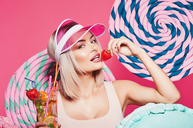 Jeune fille aux cheveux blonds portant haut et chapeau rose debout avec d'énormes sucettes sucrées à rose