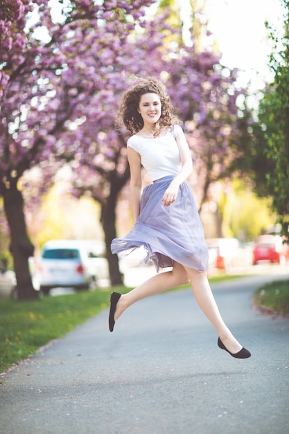 Jeune fille aux cheveux bouclés dans un t-shirt blanc et jupe violette dansant et sautant sous les arbres de sakura en fleurs