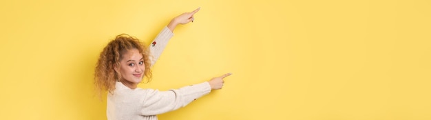 Une jeune fille aux cheveux bouclés pointe d'un geste sur un fond de studio