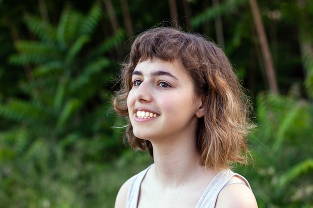 Jeune fille aux cheveux courts et bouclés souriante avec les mains près du visage. Jolie fille adolescente à l'extérieur