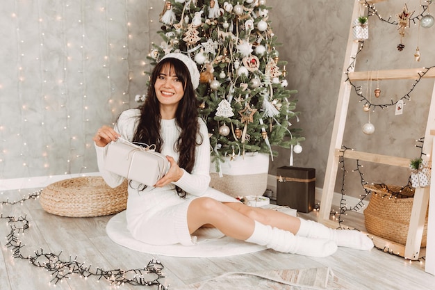 Une jeune fille aux cheveux longs noirs et vêtue d'un bonnet blanc est assise près d'un arbre de Noël et tient un cadeau dans ses mains.