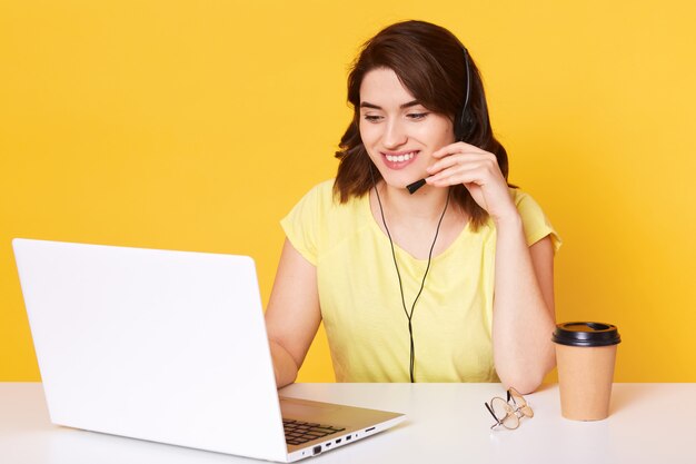 jeune fille aux cheveux noirs dans les écouteurs avec microphone est assis à table avec ordinateur portable, travaille sur le clavier et parle avec le client, boire du café, a un sourire agréable, vêtu d'un t-shirt jaune.