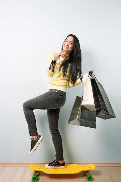 Une jeune fille aux cheveux noirs vêtue d'un chemisier blanc et jaune et d'un jean gris se dresse sur la planche à roulettes jaune tenant beaucoup de sacs après avoir fait du shopping sur le fond blanc.