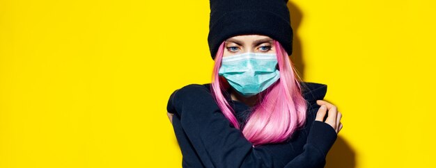 Jeune fille aux cheveux roses et aux yeux bleus, portant un masque de grippe médicale, vêtue d'un pull à capuche noir et bonnet sur le mur jaune.