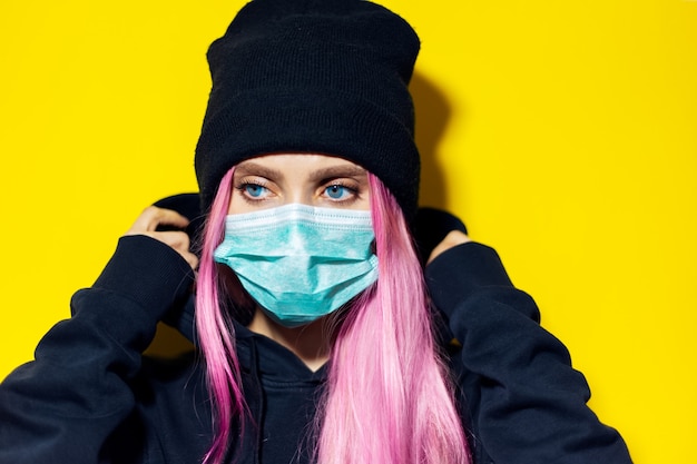 Jeune fille aux cheveux roses et aux yeux bleus, portant un masque de grippe médicale, vêtue d'un pull à capuche noir et bonnet sur le mur jaune.