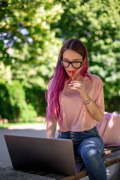 Jeune fille aux cheveux roses étudie dans le parc du printemps, assise sur le banc en bois et naviguant sur son ordinateur portable