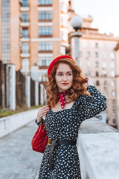 Une jeune fille aux cheveux rouges dans une robe noire et un béret rouge dans le contexte de la ville