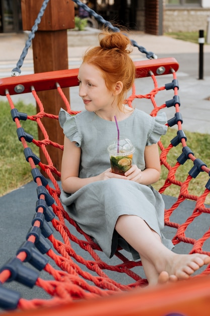 Une jeune fille aux cheveux roux est assise dans un hamac avec un verre de limonade.