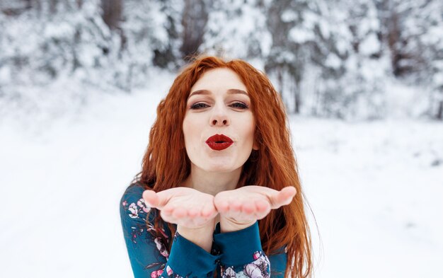 Jeune fille aux longs cheveux roux dans une robe d'été sur un paysage d'hiver. Souffler un baiser