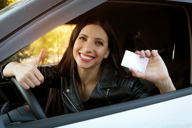 Photo jeune fille avec un beau sourire assis dans une voiture blanche montrant une carte blanche vide et un signe de la main. la femme a le permis de conduire et est très heureuse. jolie fille au volant à l'intérieur de la voiture est excitée.