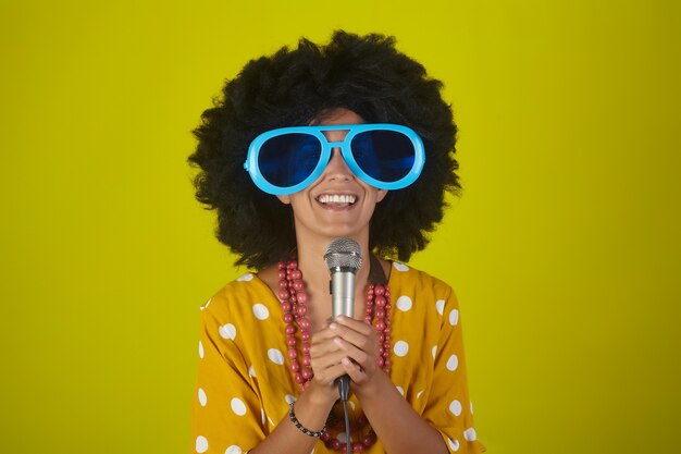 Jeune fille belle et souriante avec une coiffure afro bouclée et des lunettes drôles chantant avec microphone sur fond jaune