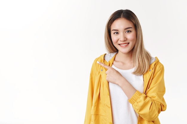 Une jeune fille blonde asiatique sincère et tendre suggère une promotion d'espace de copie cool pointant l'index gauche souriant à pleines dents attentionné conversation douce discuter d'une publicité géniale, mur blanc