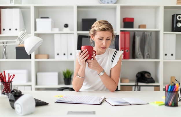 Une jeune fille blonde assise à un bureau d’ordinateur dans le bureau tient un crayon et une tasse rouge à la main