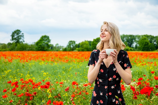 Jeune fille blonde en belle robe avec une tasse de café dans le champ de coquelicots en été