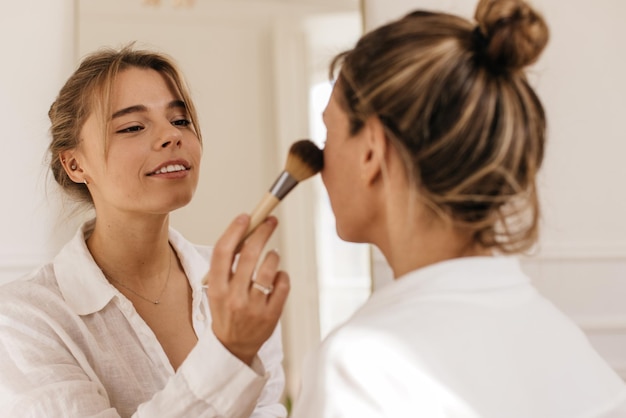 Une jeune fille blonde caucasienne applique du fard à joues sur le visage du client avec une brosse en studio léger. Concept beauté et mode