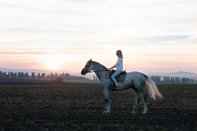 Jeune fille blonde à cheval sur le terrain pendant le coucher du soleil