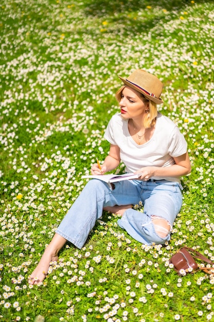 Une jeune fille blonde dans un chapeau écrivant des poèmes dans un cahier au printemps dans un parc de la nature de la ville assis sur l'herbe à côté de marguerites