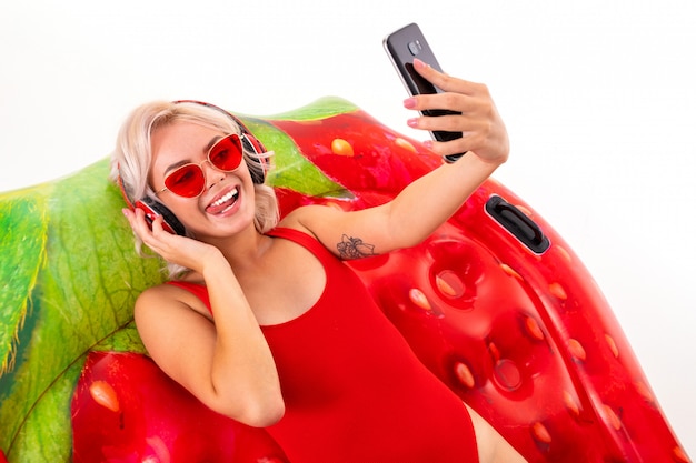 jeune fille blonde dans un maillot de bain rouge et des lunettes de soleil assis sur un matelas de natation et écouter de la musique sur un casque et tenant un téléphone dans ses mains